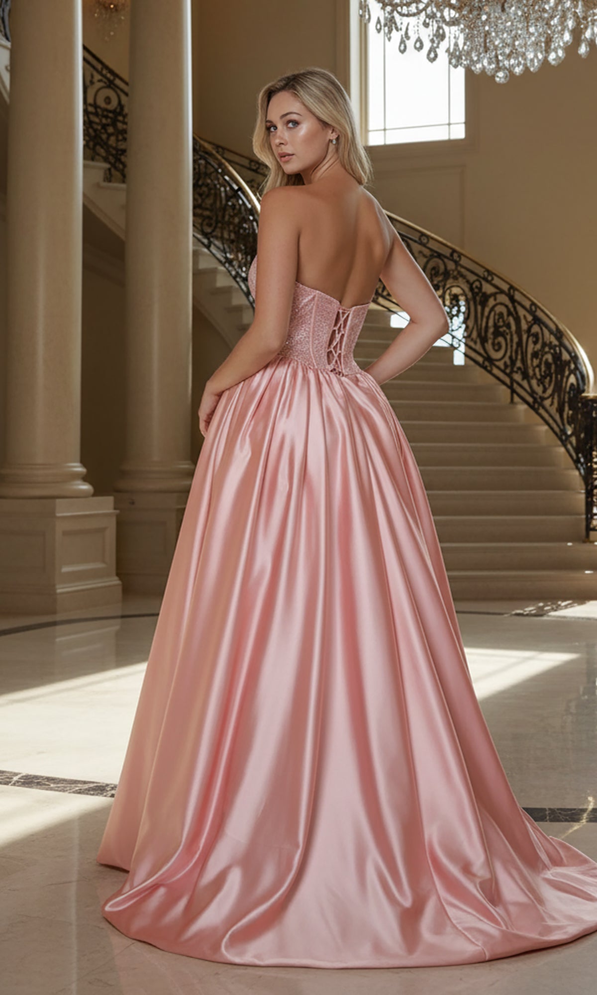 A woman wearing the Long Prom Dress: Poly USA 25156, featuring a strapless rhinestone bodice, stands in an elegant foyer with a grand staircase and sparkling chandelier adding to the glamour.
